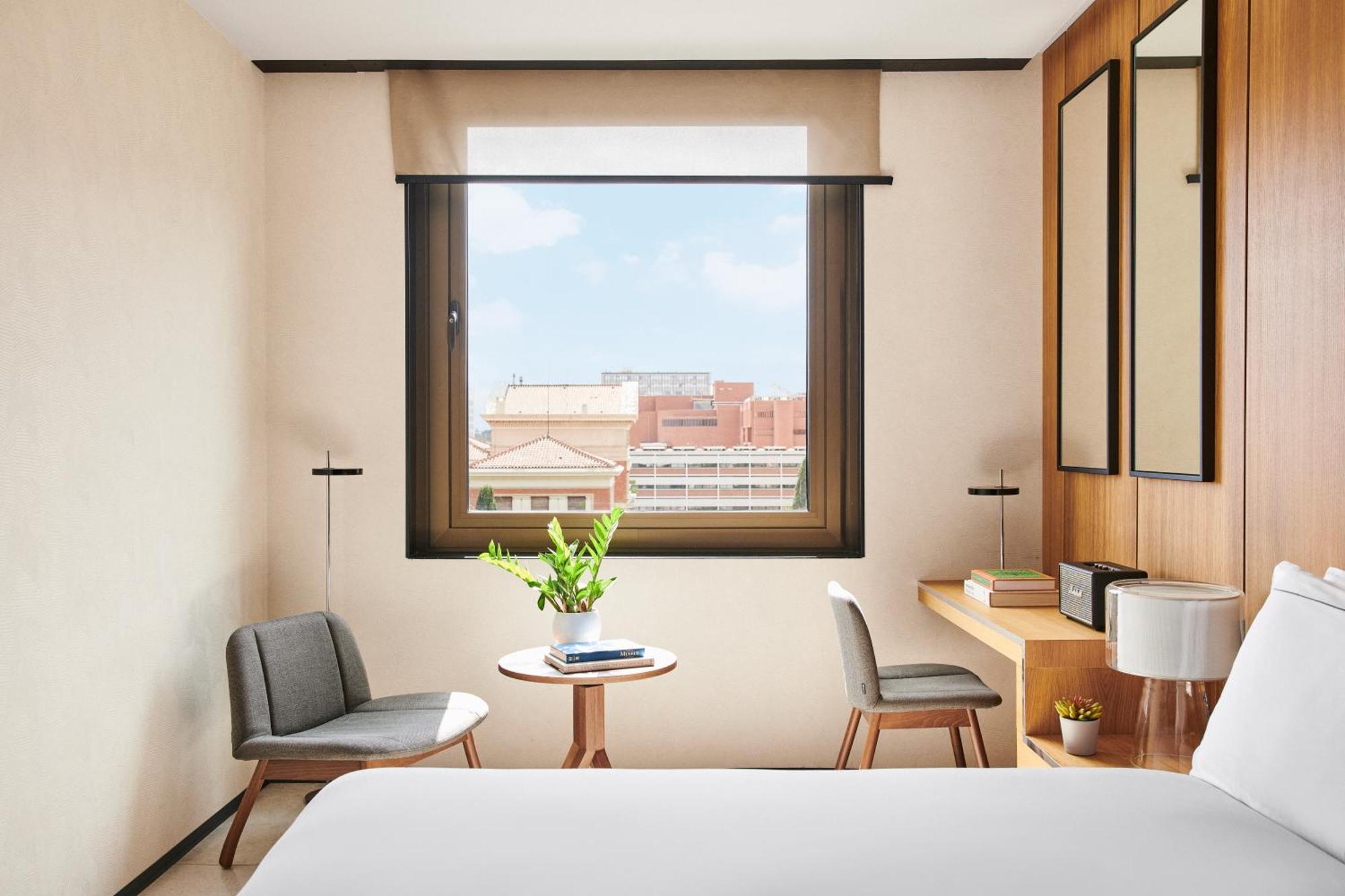 巴塞罗那索菲亚酒店-凯悦无限精选 外观 照片 The photo depicts a modern hotel room with a cozy and minimalist design. There's a large window allowing natural light to enter, showcasing a view of the outside cityscape. In the room, two chairs are positioned near a small round table, which has a 