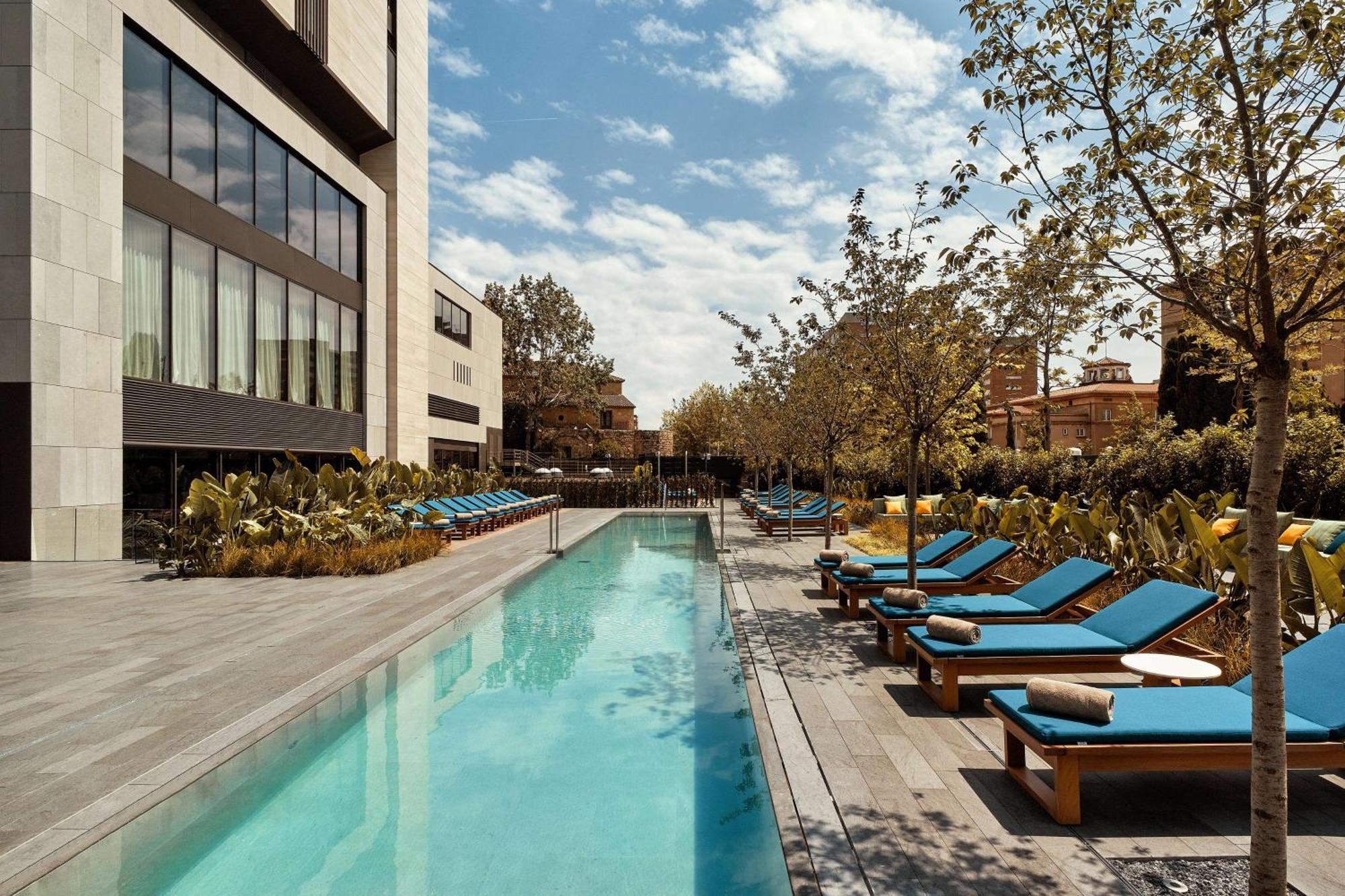 巴塞罗那索菲亚酒店-凯悦无限精选 外观 照片 The photo showcases a modern outdoor pool area. In the foreground, there is a long, rectangular swimming pool with clear blue water. Surrounding the pool are lounge chairs with bright blue cushions, arranged neatly along both sides. The area features